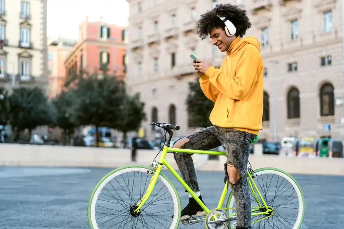 Junger Mann mit AirPods auf einem Fahrrad, der auf sein Handy schaut. Darf man mit AirPods Fahrrad fahren? Ja, aber Vorsicht!