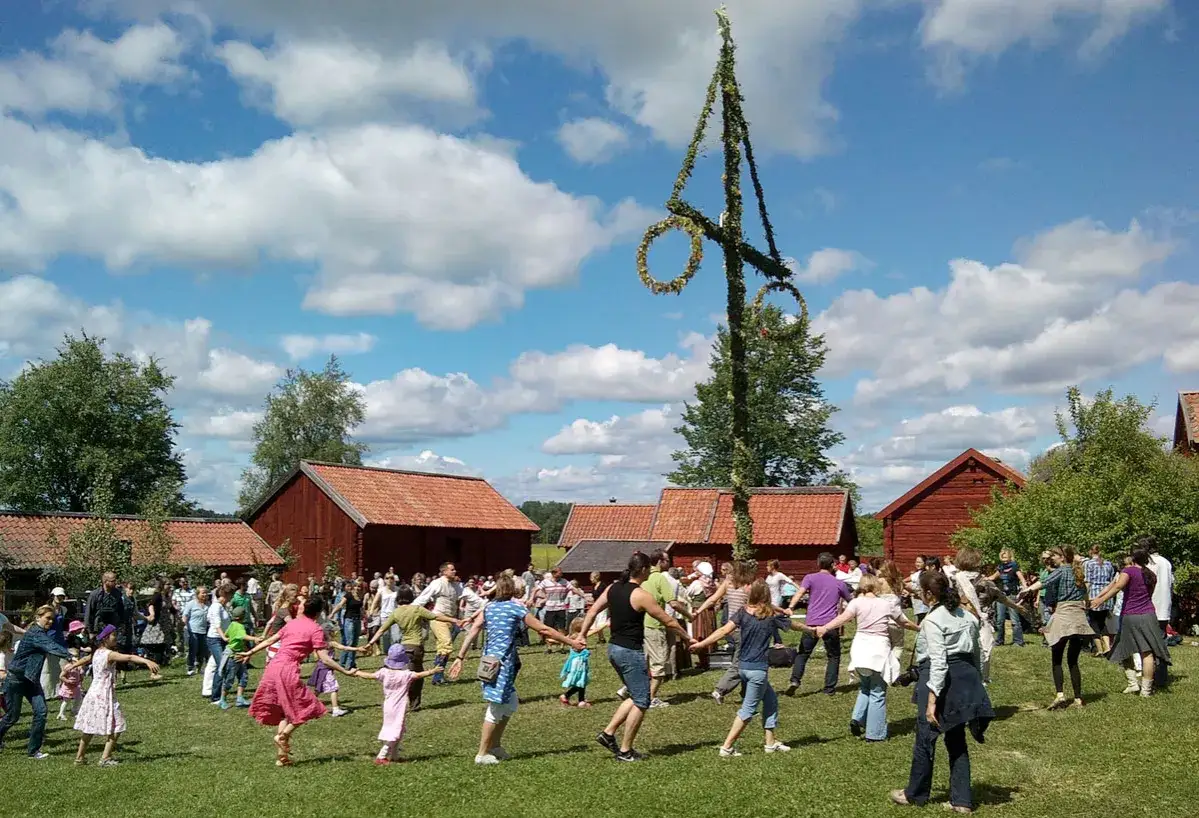 Midsommarstångens betydelse firas med ringdans runt den lövade stången. Många människor samlas på en grönskande äng, omgivna av röda hus under en blå himmel med vita moln.