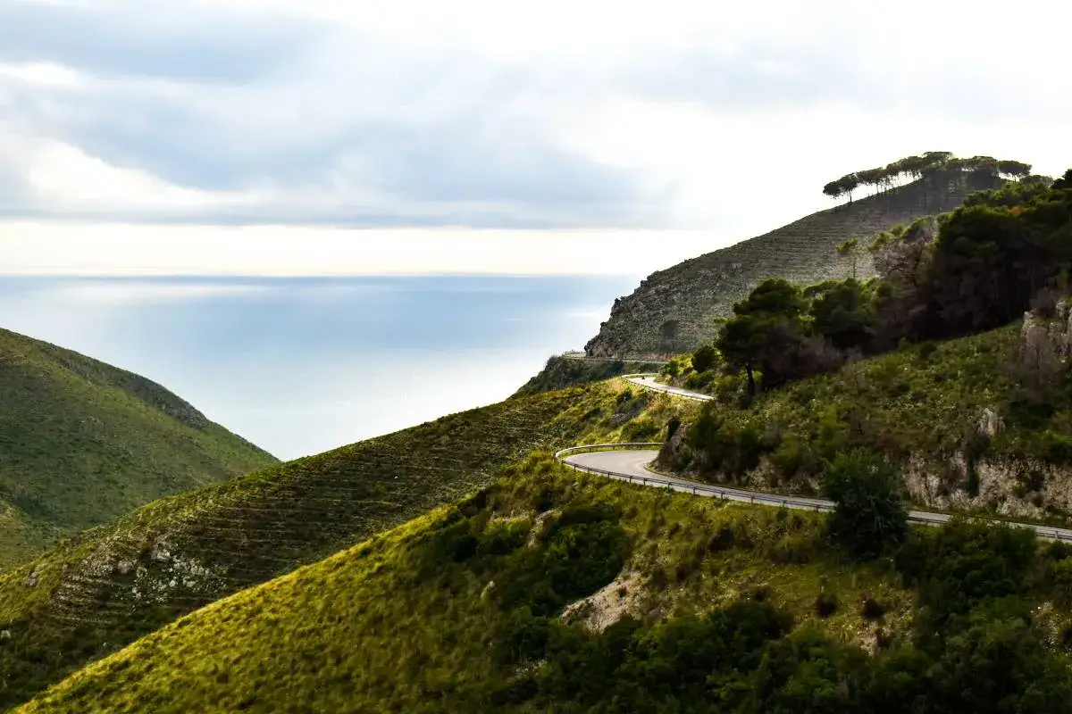 Strada tortuosa lungo la costa, ideale per un **via francigena in bici percorso** panoramico. Mare azzurro e colline verdi.