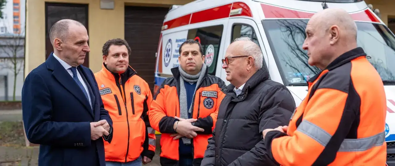 Dyskusja o tym, czy kierowca karetki musi być ratownikiem, z udziałem mężczyzn w kurtkach ratowniczych i garniturze, na tle karetki.