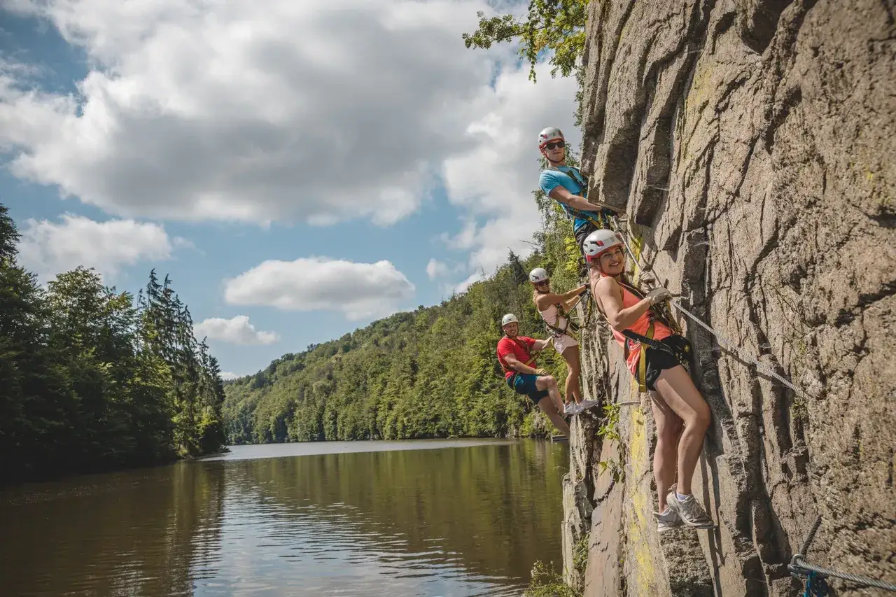 Grupa przyjaciół pokonuje via ferrata w Czechach, wspinając się po skalnej ścianie nad malowniczym jeziorem.