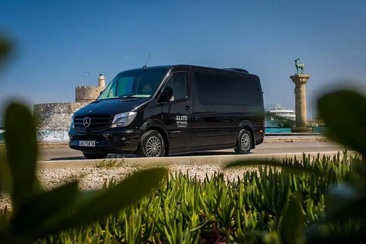 Czarny van Elite VIP Travel czeka na pasażerów przy porcie na Rodos. W tle widać zabytkową latarnię i statek wycieczkowy.