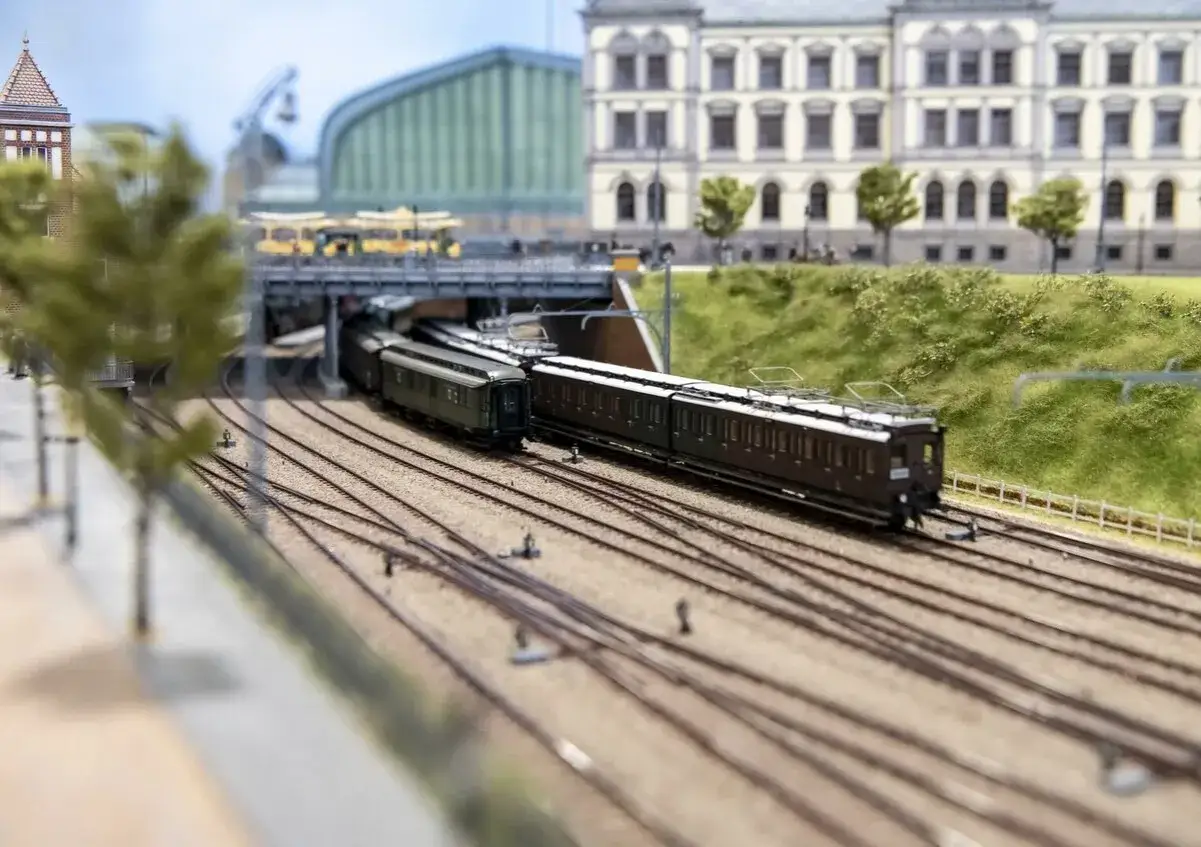 Detailreiches Dortmund Modellbau-Bahnhof mit Zügen unter einer Brücke, im Hintergrund ein Gebäude und grüne Hügel.