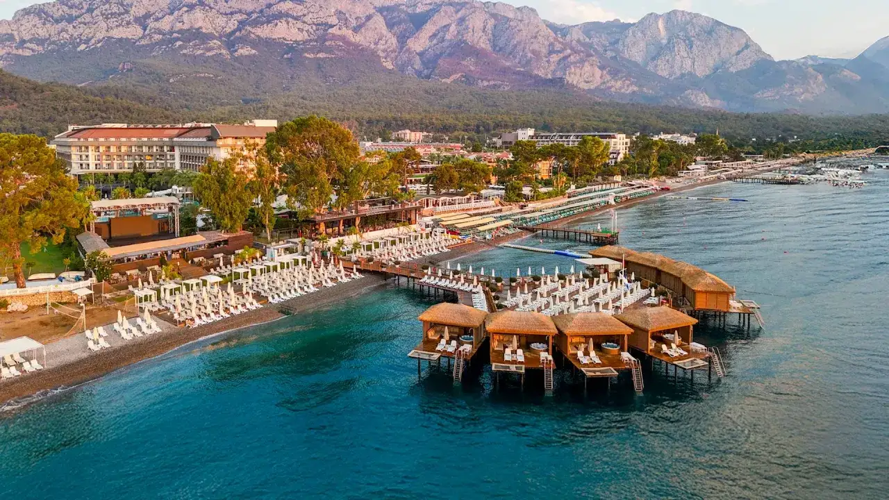 Malowniczy widok na Baia Salima Kemer z luksusowymi domkami na wodzie, plażą i górami w tle.