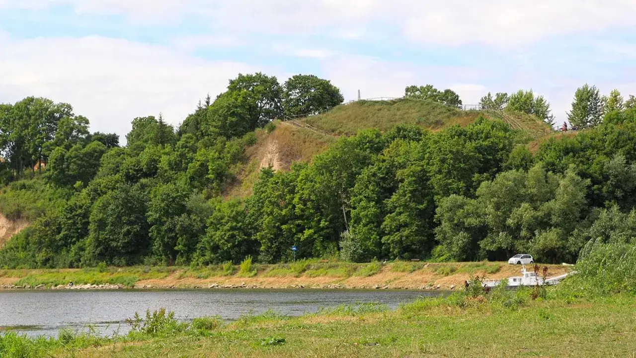 Zielona skarpa nad rzeką, na szczycie której widać ścieżki i ogrodzenie. To malownicza góra zamkowa w Drohiczynie.