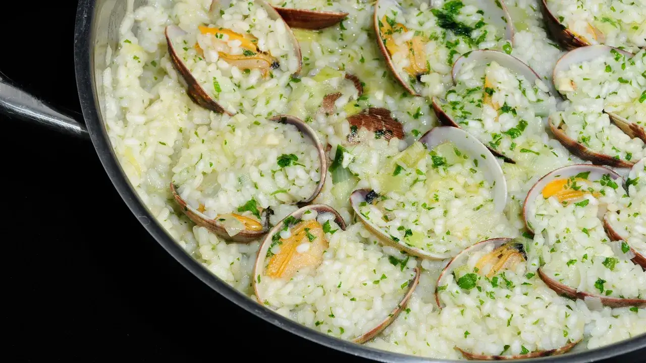 Arroz cremoso con almejas frescas y perejil picado, servido en una sart&eacute;n met&aacute;lica. Un plato delicioso y lleno de sabor marino.