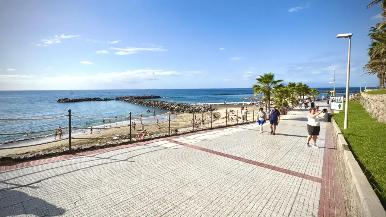 Gente pasea por el paseo marítimo de la caleta de Adeje, con la playa y el mar azul de fondo.