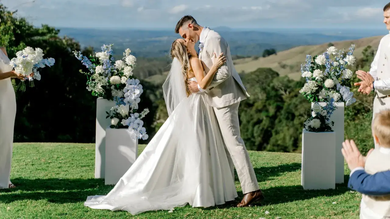 Ein Brautpaar küsst sich bei einer modernen Hochzeit im Freien. Der Bräutigam trägt einen hellen Anzug, die Braut ein weißes Kleid. Blumenarrangements säumen den Weg.