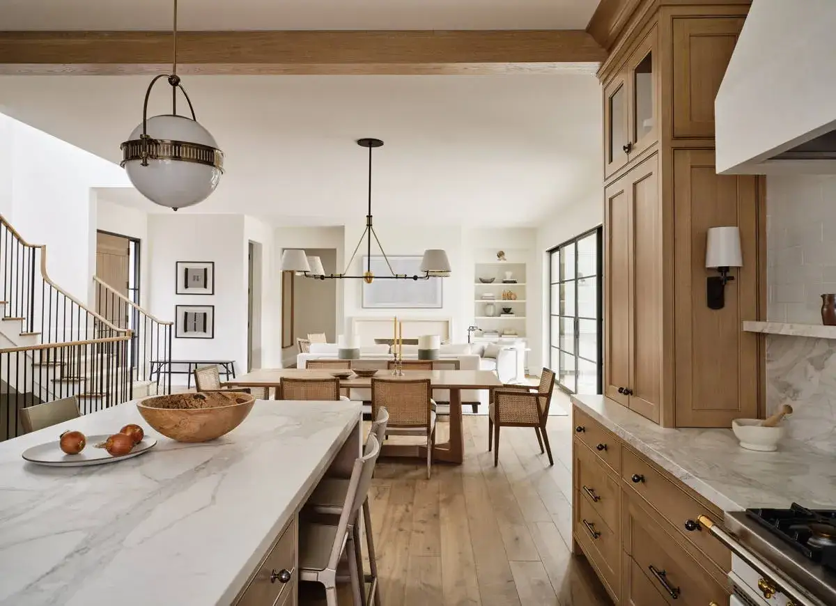 A spacious, open-plan living area featuring a natural wood kitchen, dining room, and living room. The kitchen island has a marble countertop and a bowl of apples.