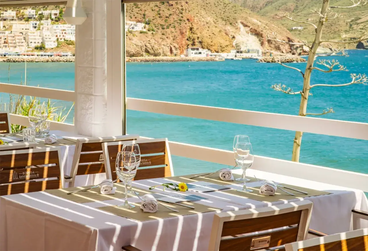 Mesas puestas en un restaurante con vistas al mar en San José. La botavara del velero se asoma entre la vegetación.