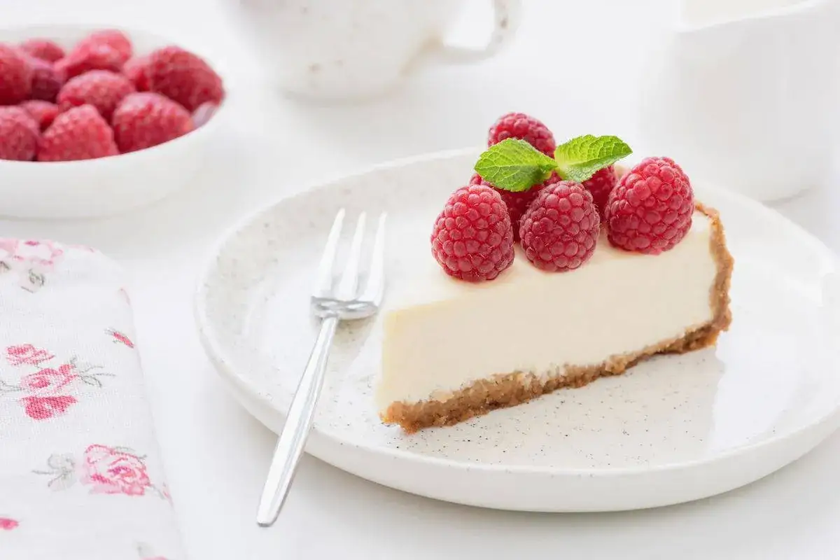 Una fetta di torta fredda allo yogurt guarnita con lamponi freschi e menta, su un piatto bianco con una forchetta.