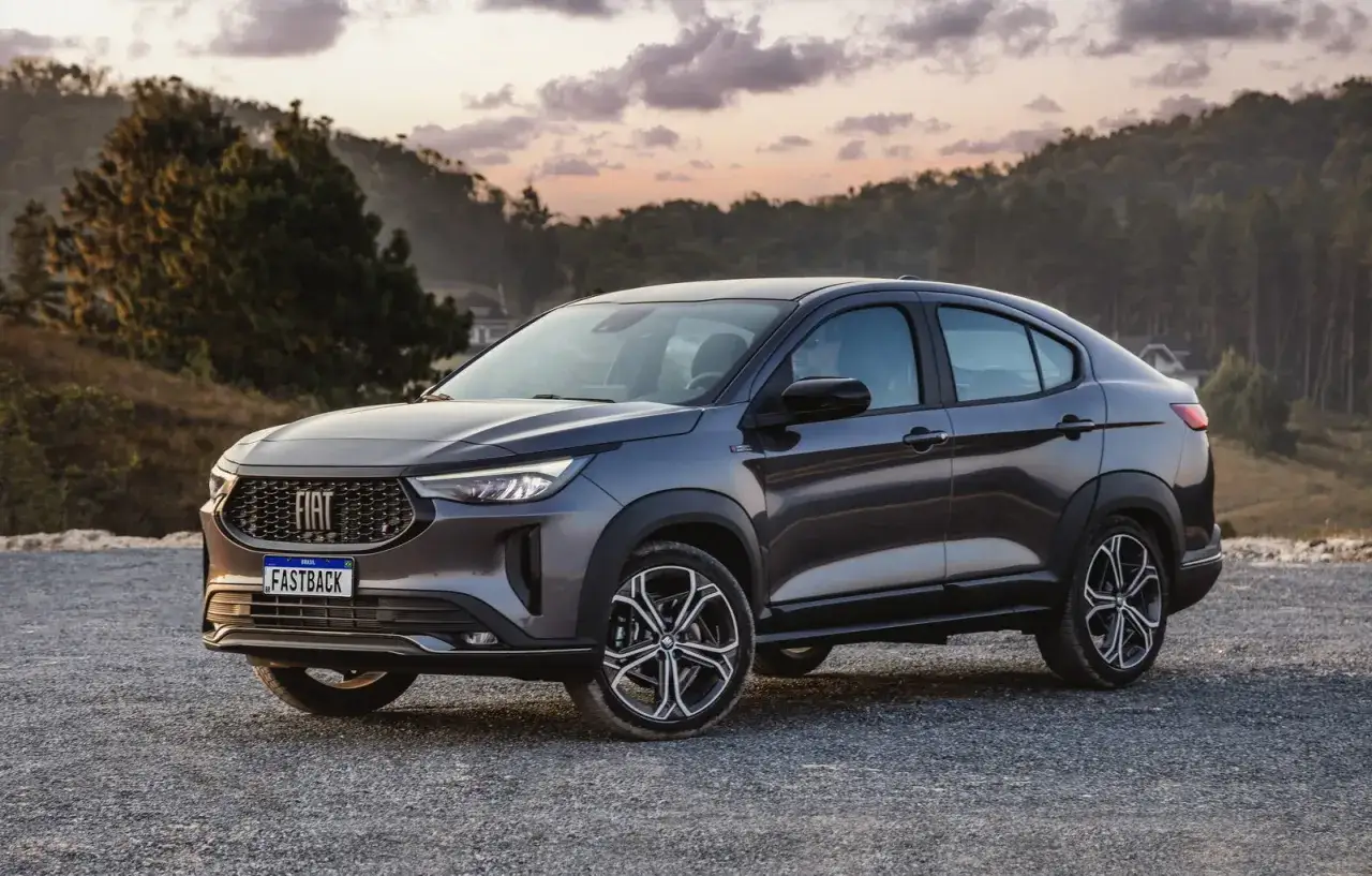 Fiat Fastback 2026, un SUV coup&eacute; grigio scuro, parcheggiato su ghiaia con colline boscose sullo sfondo al tramonto.
