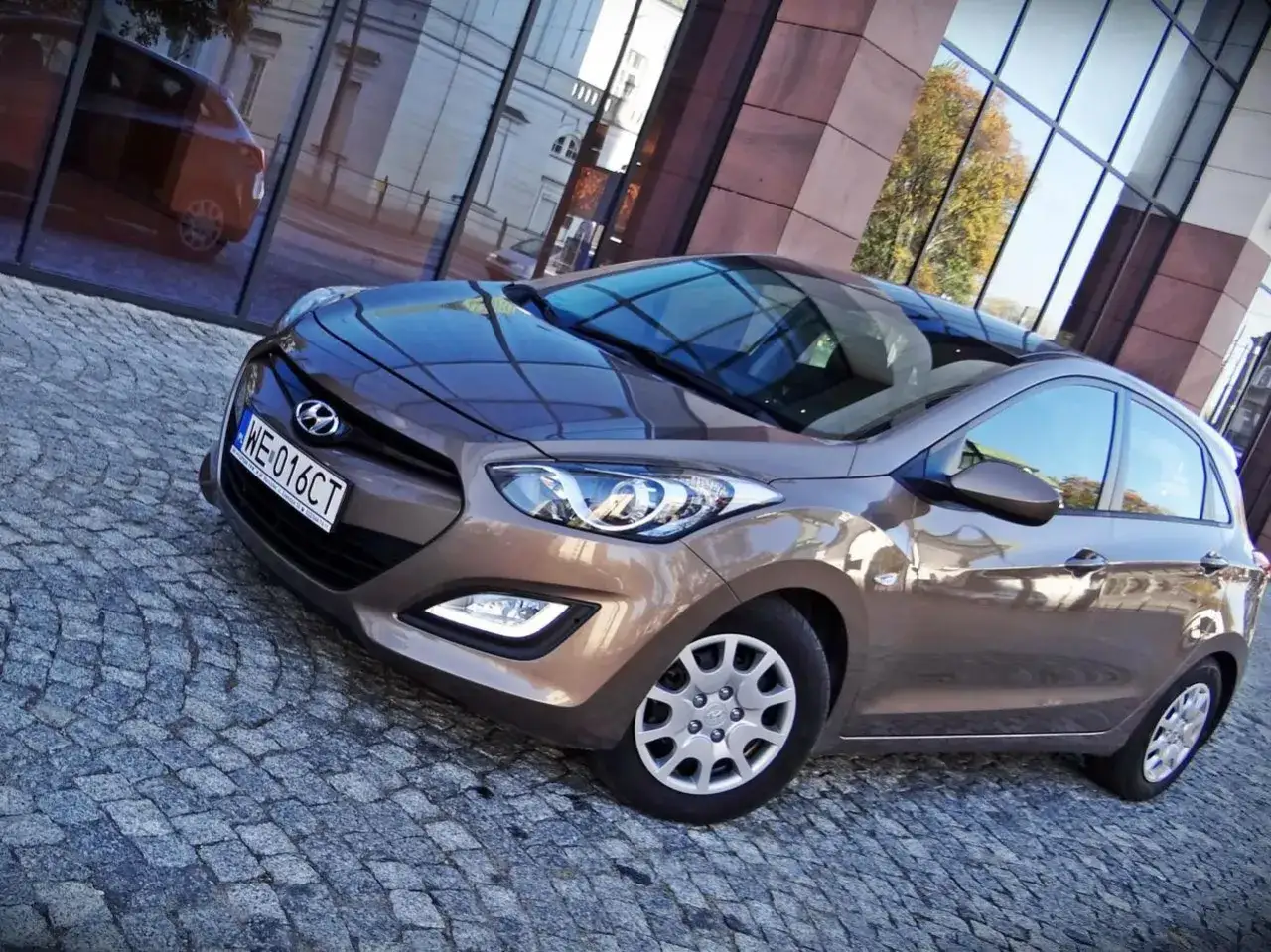 Brązowy Hyundai i30, jedna z popularnych generacji, zaparkowany na brukowanej ulicy przed nowoczesnym budynkiem.