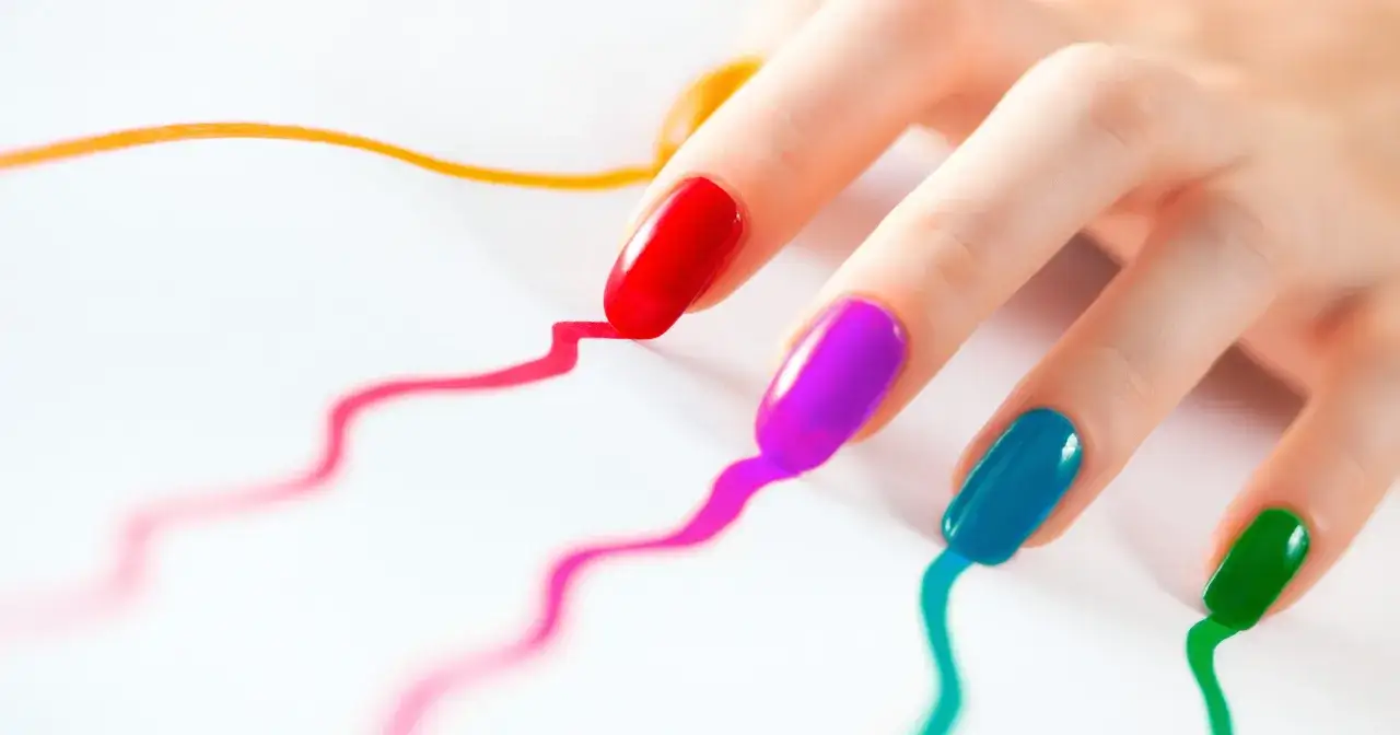 Dłoń z kolorowymi paznokciami (czerwony, fioletowy, niebieski, zielony) na białym tle, jakby malowała kolorowe linie. Idealny manicure japoński w ciąży.