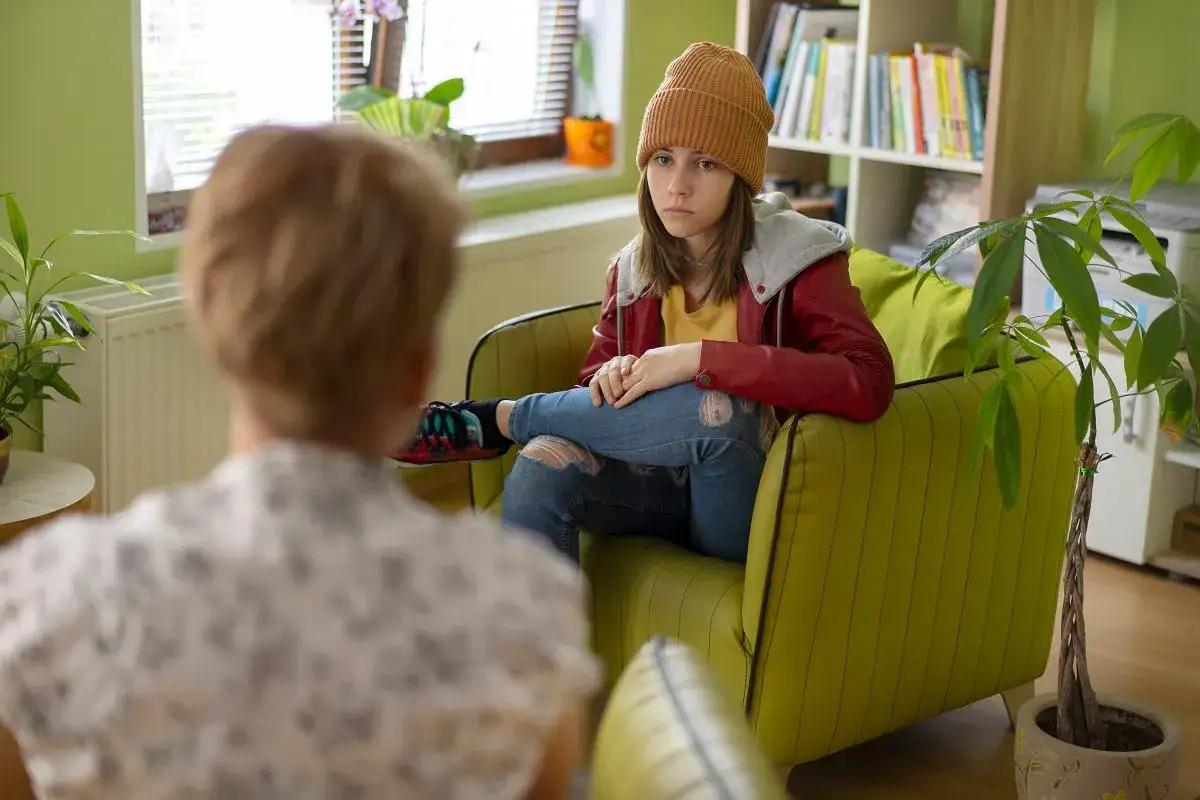 Nastolatka w beżowej czapce i czerwonej kurtce siedzi na kanapie, rozmawiając z terapeutką. Dyskutują o tym, od ilu lat można pracować.