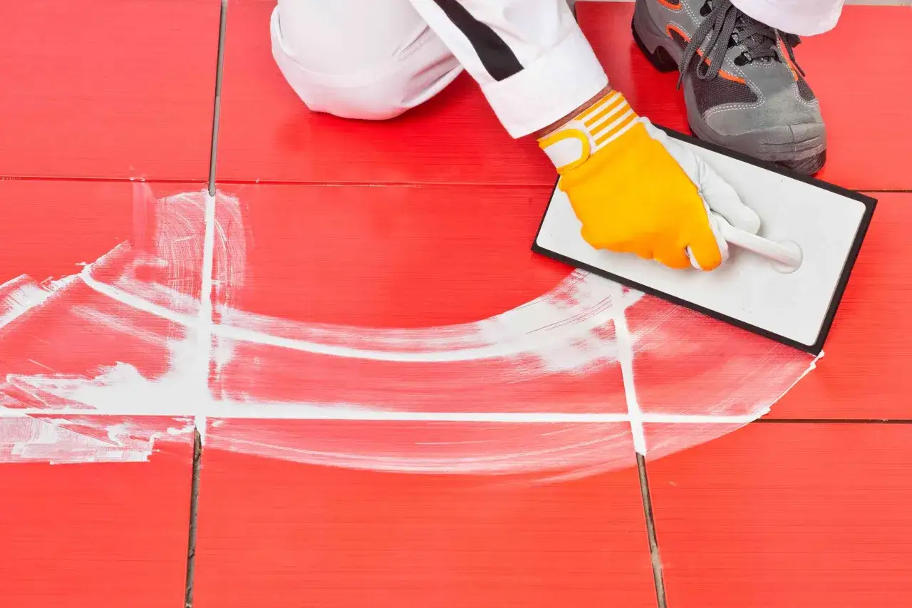 Man con guantes amarillos aplica lechada blanca a azulejos rojos con una llana.