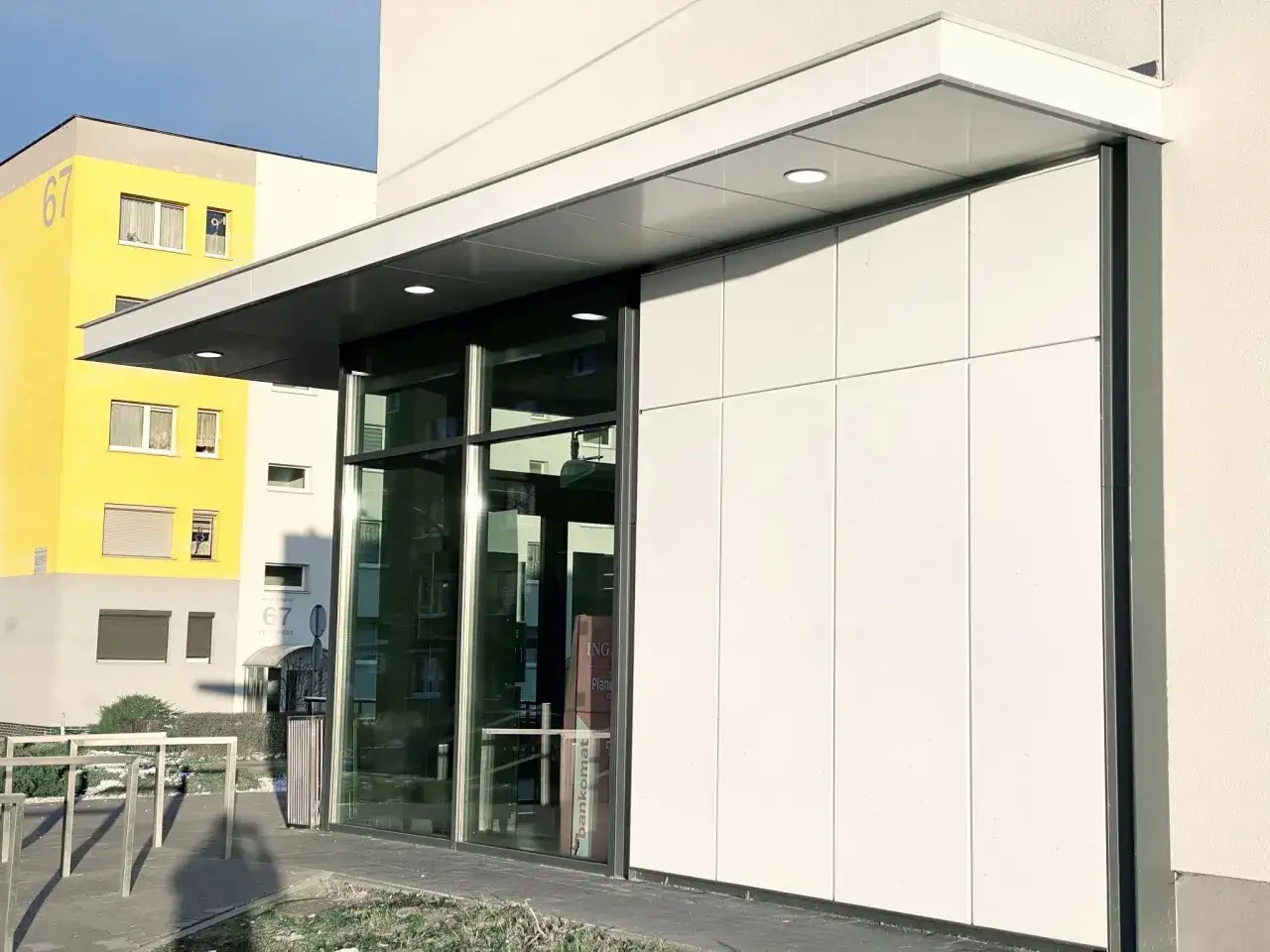 Nowoczesne wejście do budynku z panelami alucobond, przeszklonymi drzwiami i żółtym budynkiem w tle.