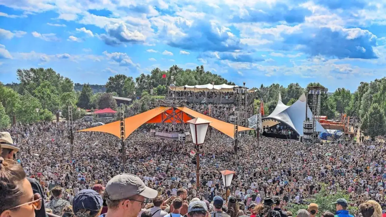 Tausende feiern auf dem Fusion Festival Ort unter blauem Himmel. Bunte Zelte und B&uuml;hnen pr&auml;gen die Szenerie.