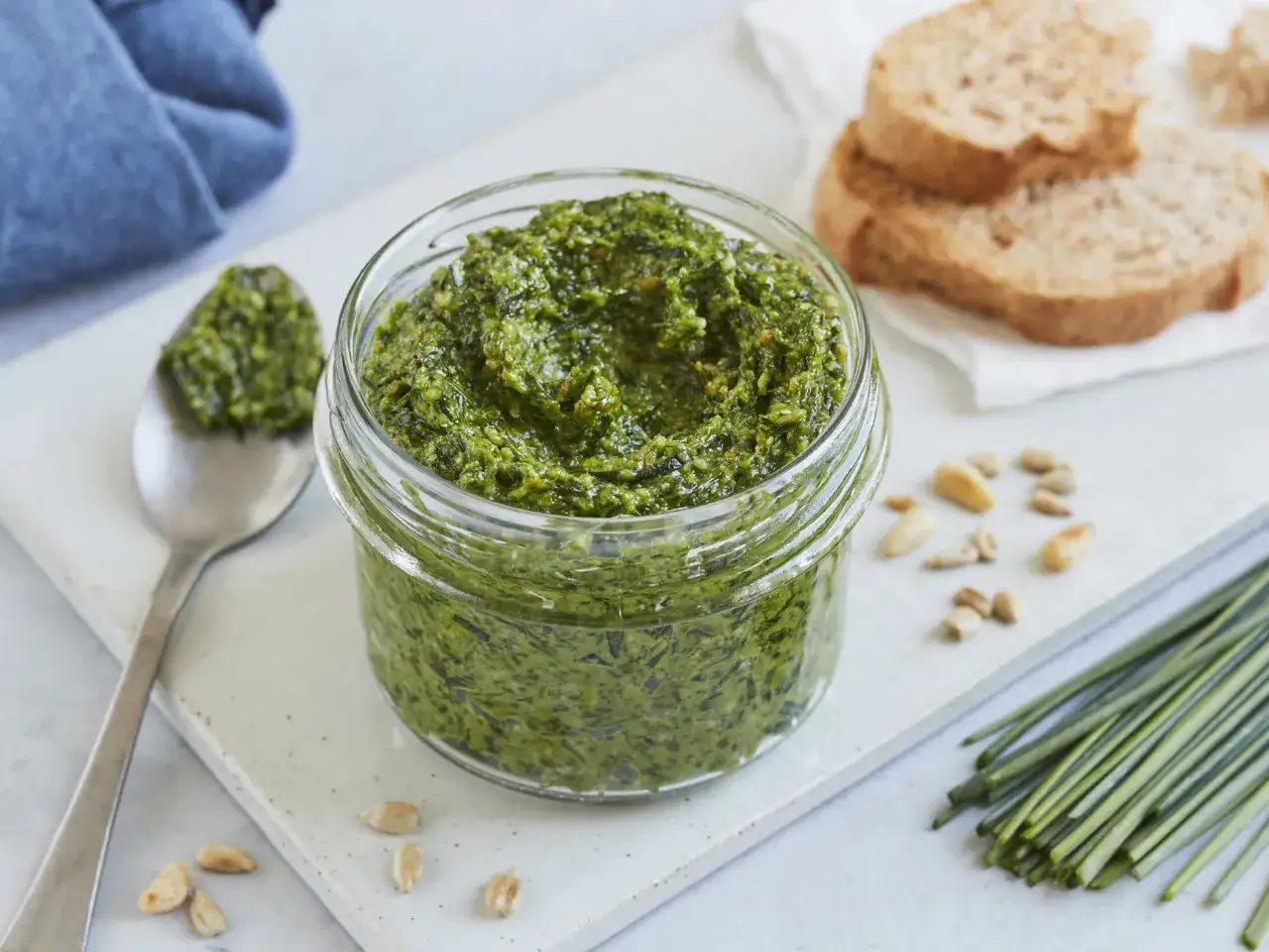 Frisches schnittlauch pesto in einem Glas, daneben eine Löffelportion, Brot und Pinienkerne.