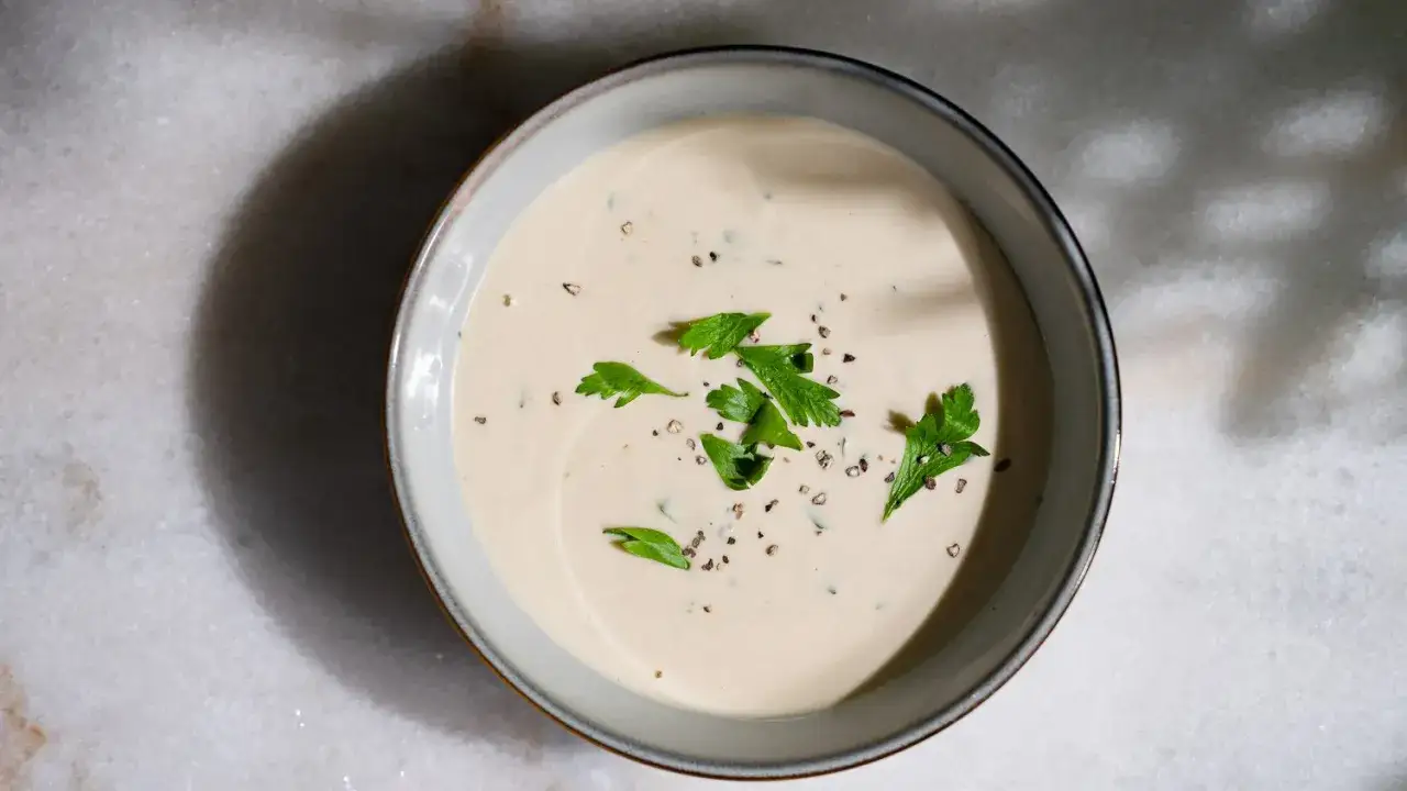 Sauce tahini cr&eacute;meuse, garnie de persil frais et de poivre noir concass&eacute;, dans un bol sur fond de marbre.