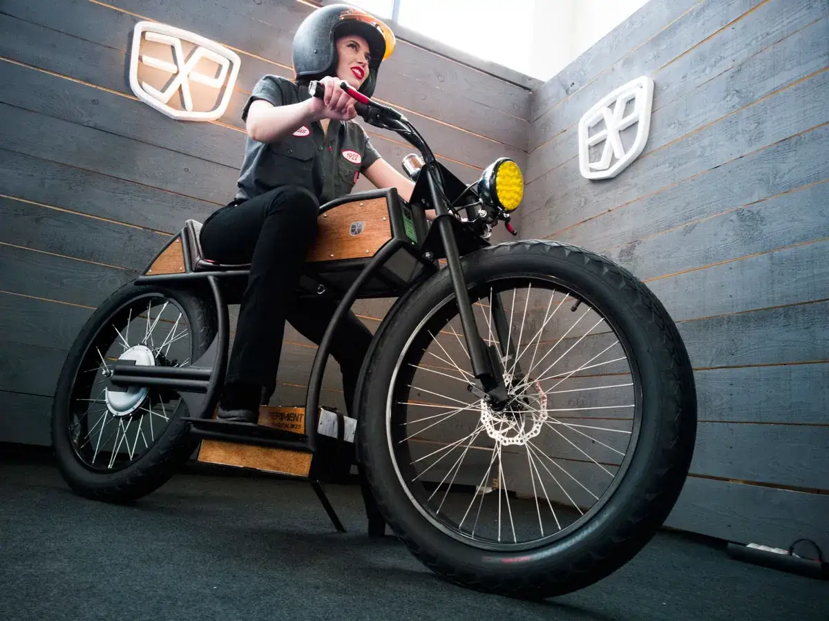Mujer con casco posa en una moto custom electrica de diseño único, con detalles en madera y rueda delantera ancha.