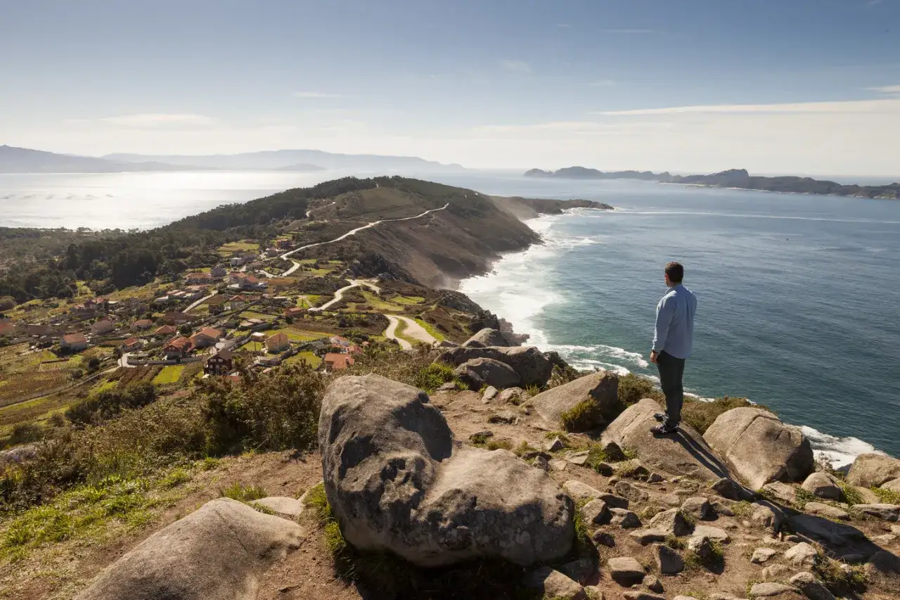 Hombre contempla el mar desde el cabo Udra, con un pueblo costero y montañas al fondo.