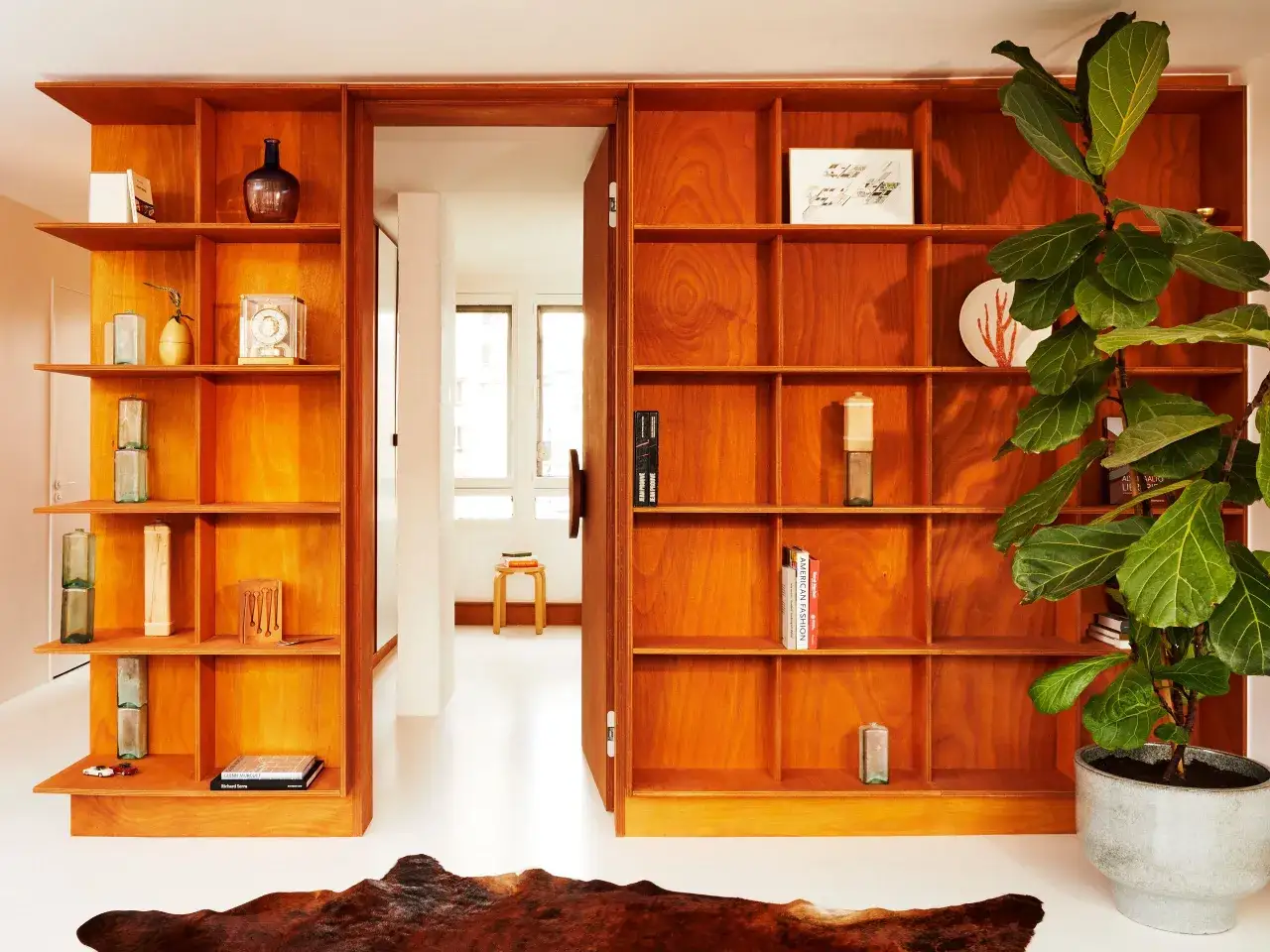 Ein offenes Bücherregal aus Holz, das einen Durchgang schafft, um aus einem Zimmer zwei zu machen. Dekoriert mit Vasen, Büchern und einer großen Zimmerpflanze.