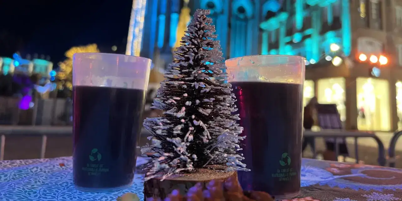 Deux verres de vin chaud sur une table décorée, avec un petit sapin au milieu. Ambiance de marché de Noël illuminé.