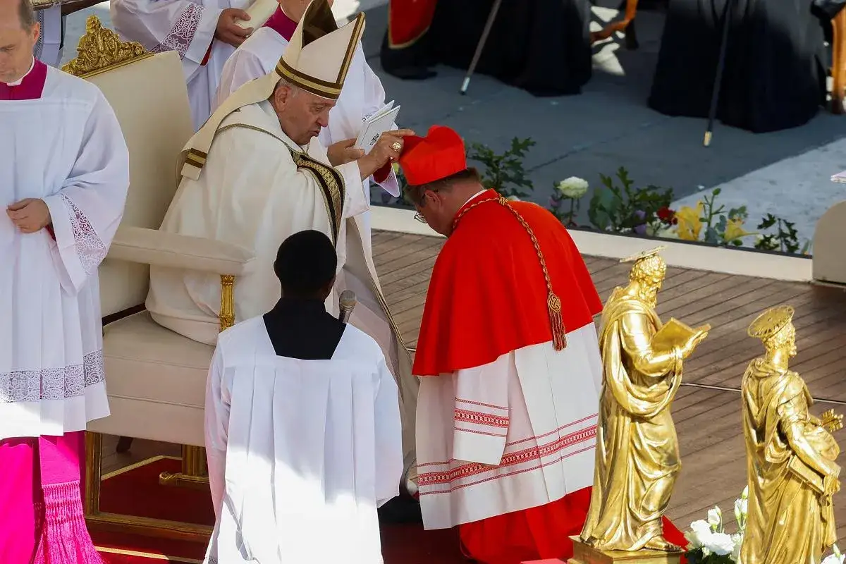Papież Franciszek w otoczeniu duchownych, w tym kardynała w czerwonej szacie. W tle złote figury świętych.
