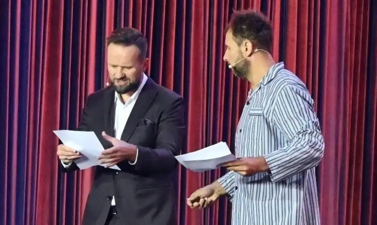 Dwóch mężczyzn, jeden w garniturze, drugi w piżamie, czyta teksty na scenie. To występ kabaretu z Kielc.
