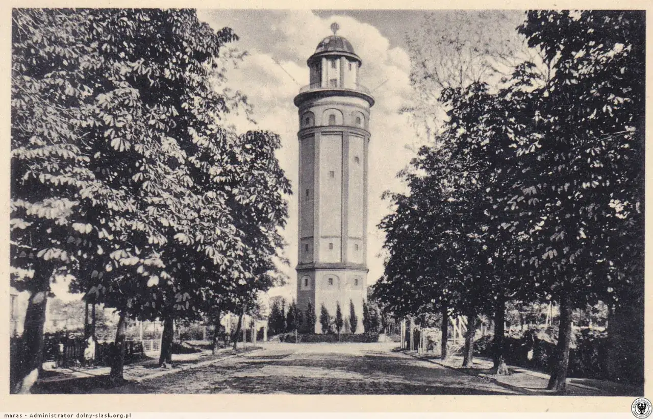 Wieża ciśnień w Karłowicach, otoczona drzewami, góruje nad parkowym krajobrazem.
