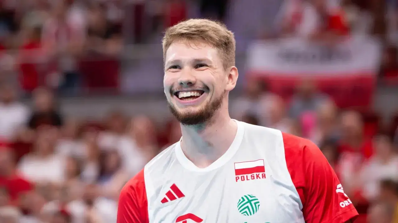 Uśmiechnięty siatkarz Jakub Kochanowski, reprezentujący Polskę, z dumą nosi biało-czerwoną koszulkę. Jego wzrost pozwala mu na skuteczną grę.