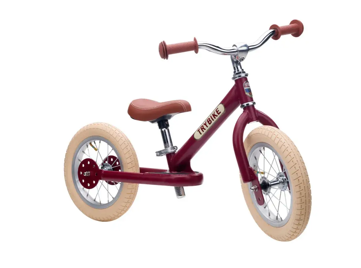 Bordowy rowerek biegowy Trybike, idealny na pierwszy rower. Ma brązowe siodełko i uchwyty, kremowe opony.