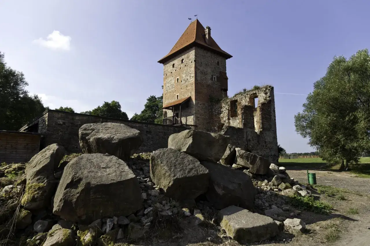 Kamienny zamek w Chudowie, ruiny i wieża z czerwoną dachem. Wokół wielkie głazy, zielone drzewa i błękitne niebo. Chudów atrakcje warte zobaczenia.
