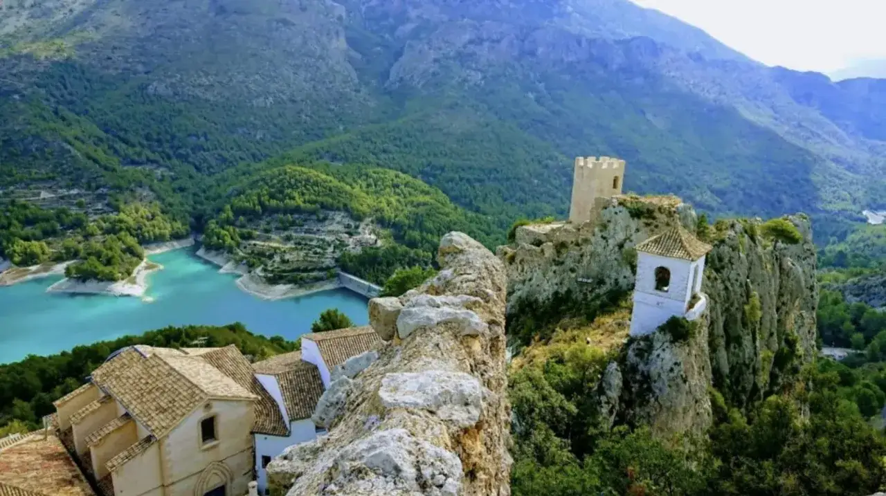 Vista panor&aacute;mica del embalse de Guadalest con su agua turquesa, rodeado de monta&ntilde;as verdes y el castillo en la cima.