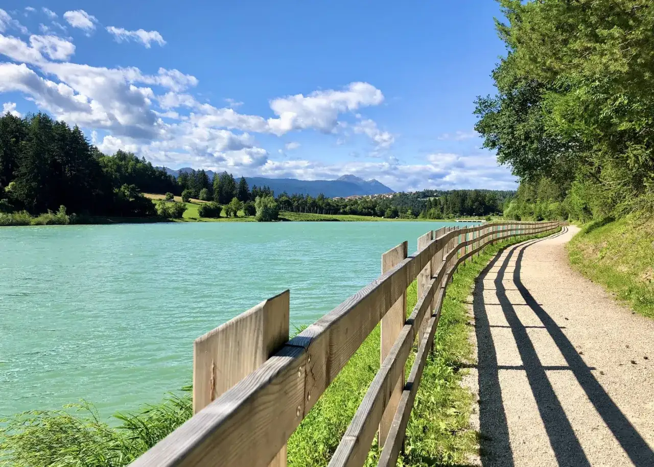 Sentiero sterrato lungo il lago di Tavon, con una staccionata in legno e montagne sullo sfondo.