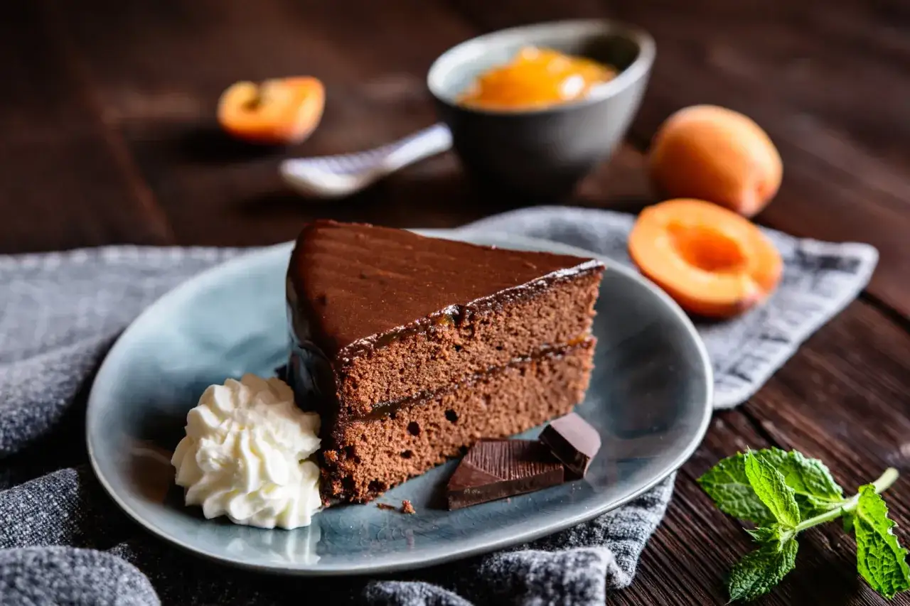 Una fetta di torta Sacher, lucida e invitante, con panna montata, albicocche e cioccolato.