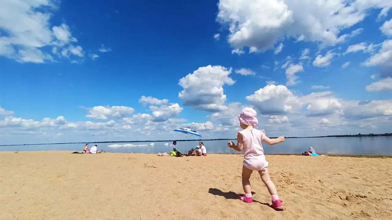 Dziewczynka w r&oacute;żowym ubranku bawi się na piaszczystej plaży. W tle widać ludzi odpoczywających pod parasolem i spokojną taflę wody. To idealny dzień na **ostrowie warckim**.