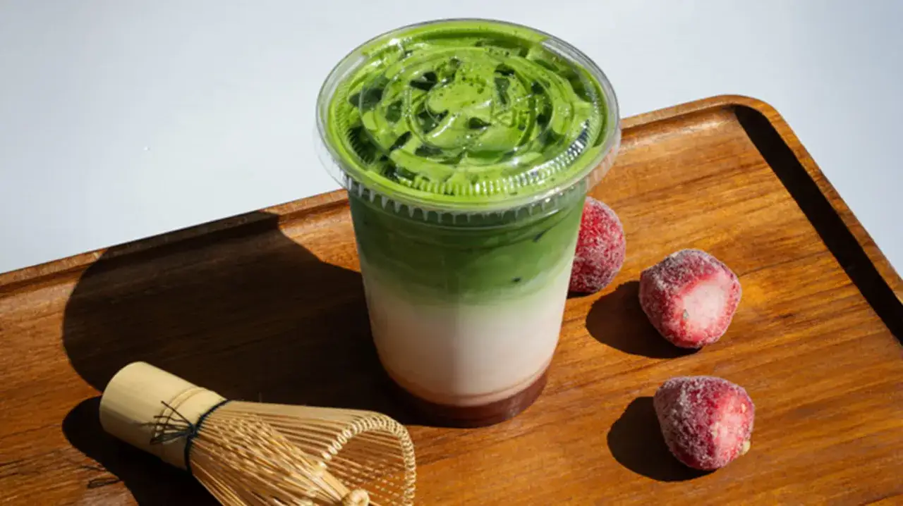En läcker, lagerdelad matcha latte i en genomskinlig mugg, med frysta jordgubbar och en bambuvisp. Matcha te nyttigt för en hälsosam start.