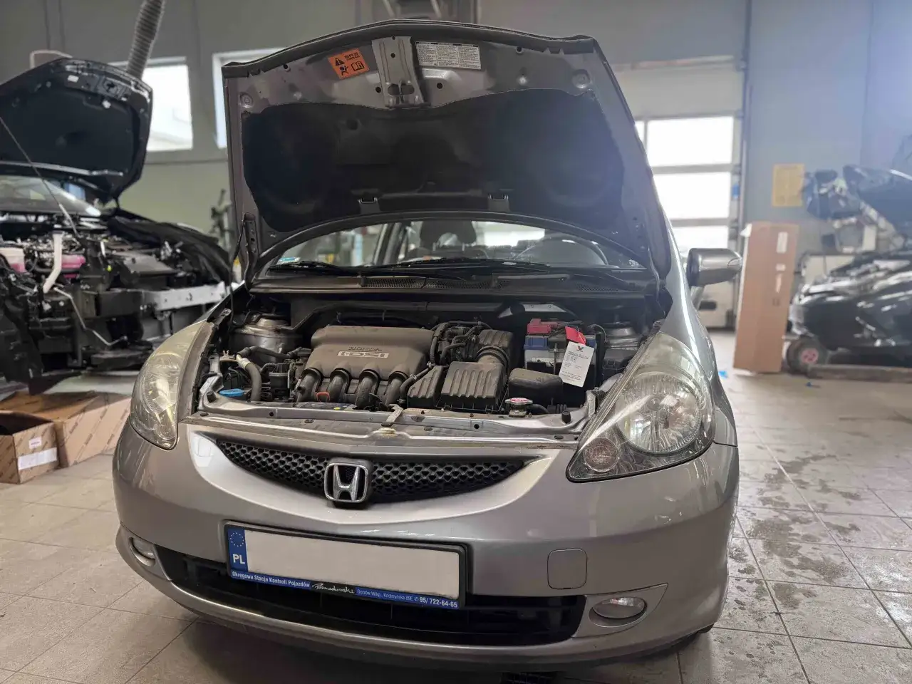 Srebrna Honda Jazz z otwartą maską, widoczny silnik i akumulator. Wymiana żarówki Honda Jazz w trakcie.