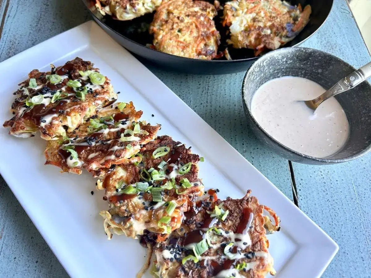 Okonomiyaki recept: Krispiga pannkakor med kål, morot och lök, toppade med sås och sesamfrön. Serveras med en krämig dippsås.
