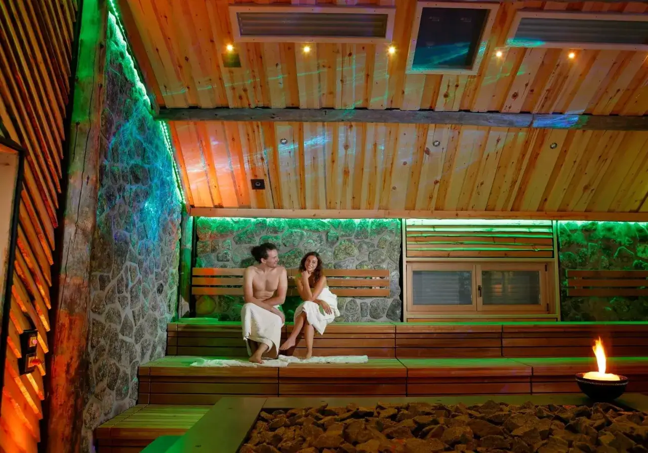 Ein Paar entspannt in einer Sauna mit H2O-Schwimmbad-Atmosph&auml;re. Gr&uuml;ne Lichter und Holz schaffen eine gem&uuml;tliche Stimmung.