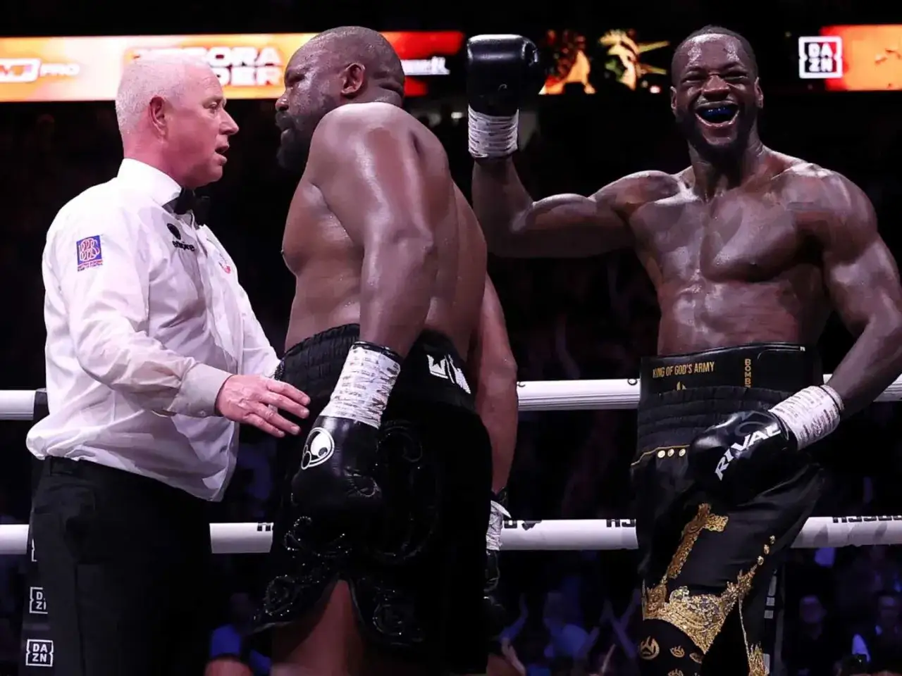 A referee stands between two boxers. One boxer, with his arm raised in victory, grins widely. The other, facing the referee, appears to be listening intently. This is a moment where you can't push in boxing.