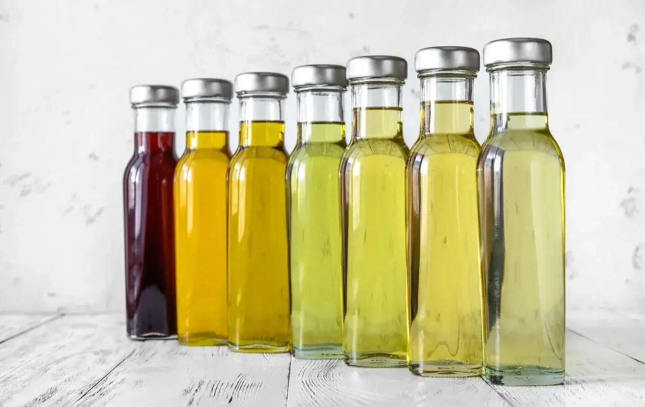 Siete botellas de vidrio con l&iacute;quidos de diferentes tonos, desde un rojo oscuro hasta varios amarillos y verdes p&aacute;lidos, mostrando la variedad del color del aceite de oliva virgen extra.