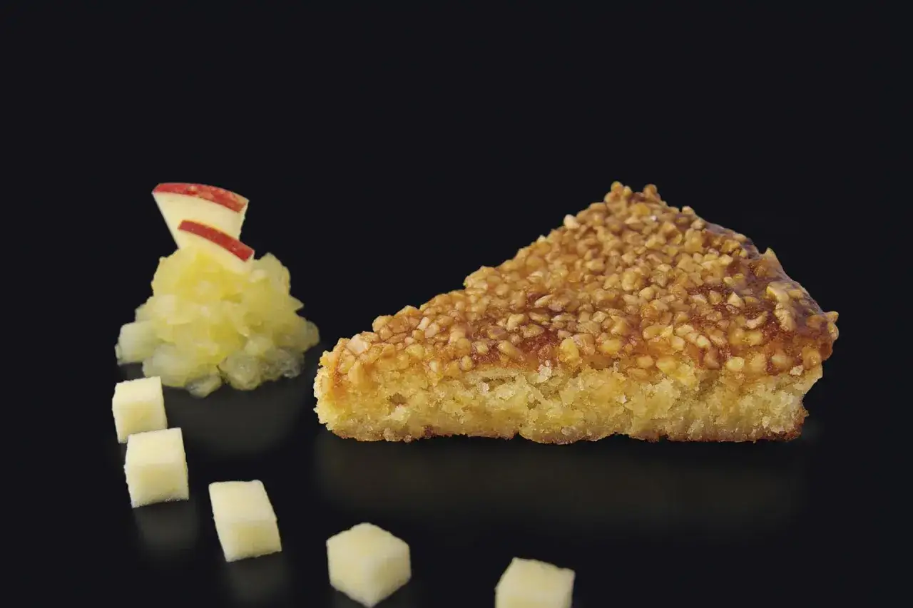 Rebanada de tarta de Mondo&ntilde;edo con cobertura crujiente de frutos secos, acompa&ntilde;ada de compota de manzana y cubos de queso.