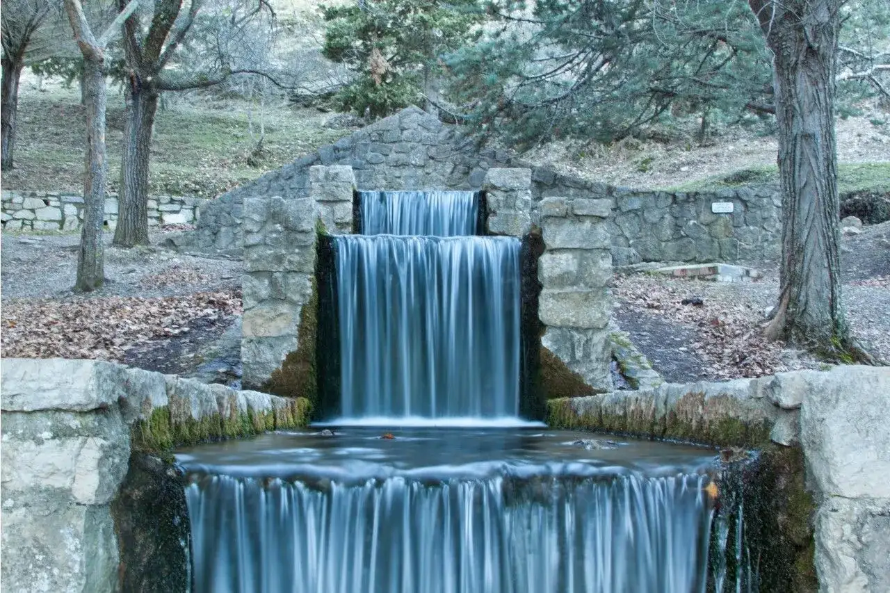 Cascada de piedra con agua sedosa cayendo en un charco del aceite. &Aacute;rboles desnudos y hojas secas rodean la escena.