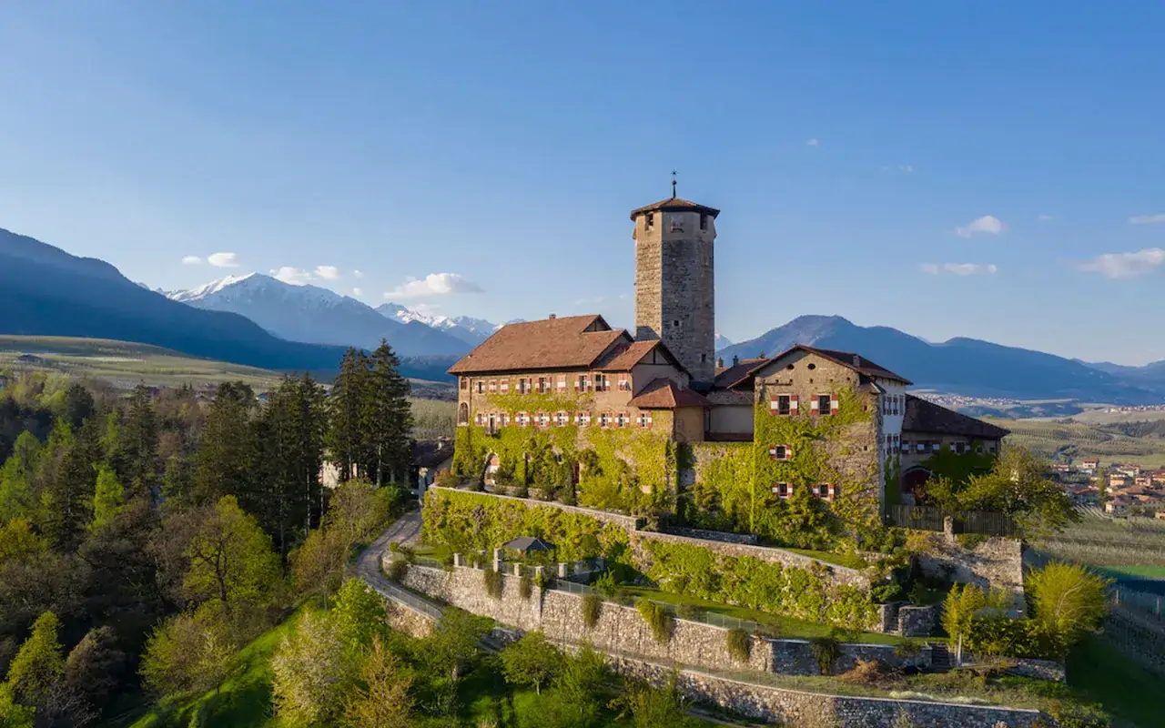 Castello medievale immerso nel verde, con torri imponenti e mura ricoperte d'edera, domina un paesaggio montuoso che ricorda i **paesi Val di Non**.