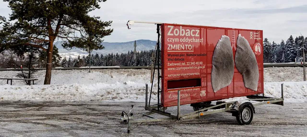 Zobacz, czym oddychasz! Zmień to! Kampania informacyjna jak zapobiegać smogowi, z grafiką płuc na czerwonej przyczepie.