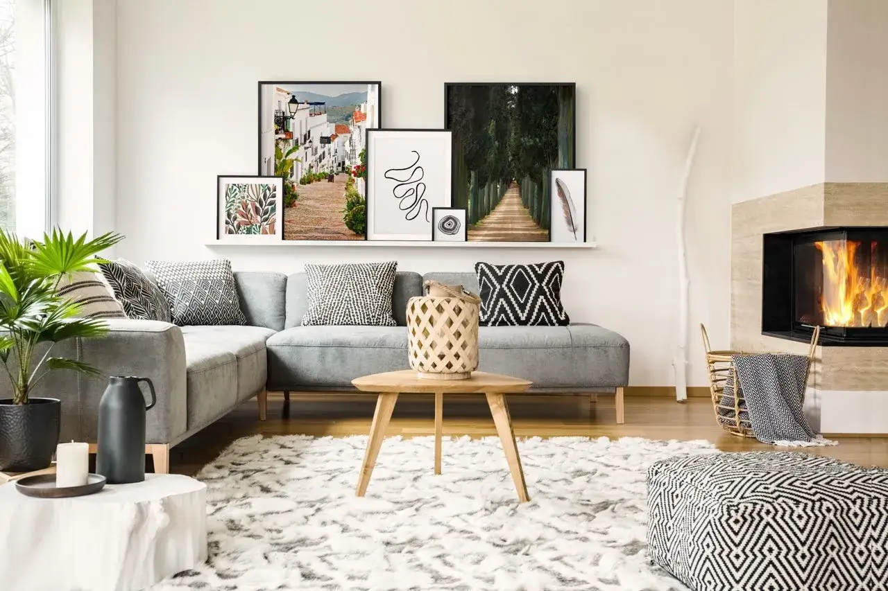 En modern soffgrupp med grå soffa och många kuddar. En hylla ovanför med flera tavlor som är fint placerade. En brasa sprakar i en öppen spis.