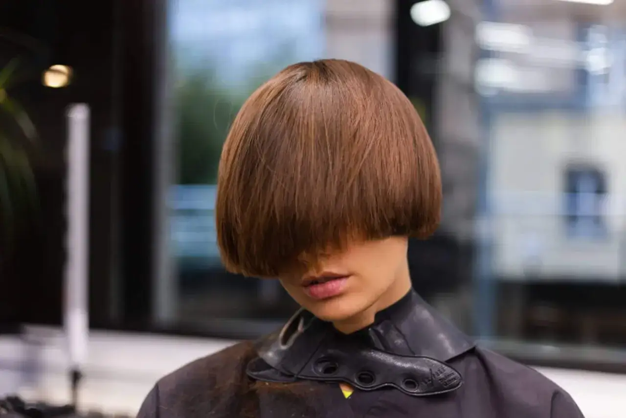 Une femme arbore une coupe au bol audacieuse, ses cheveux châtains cachant son regard. Le col noir de sa tenue ajoute une touche moderne.